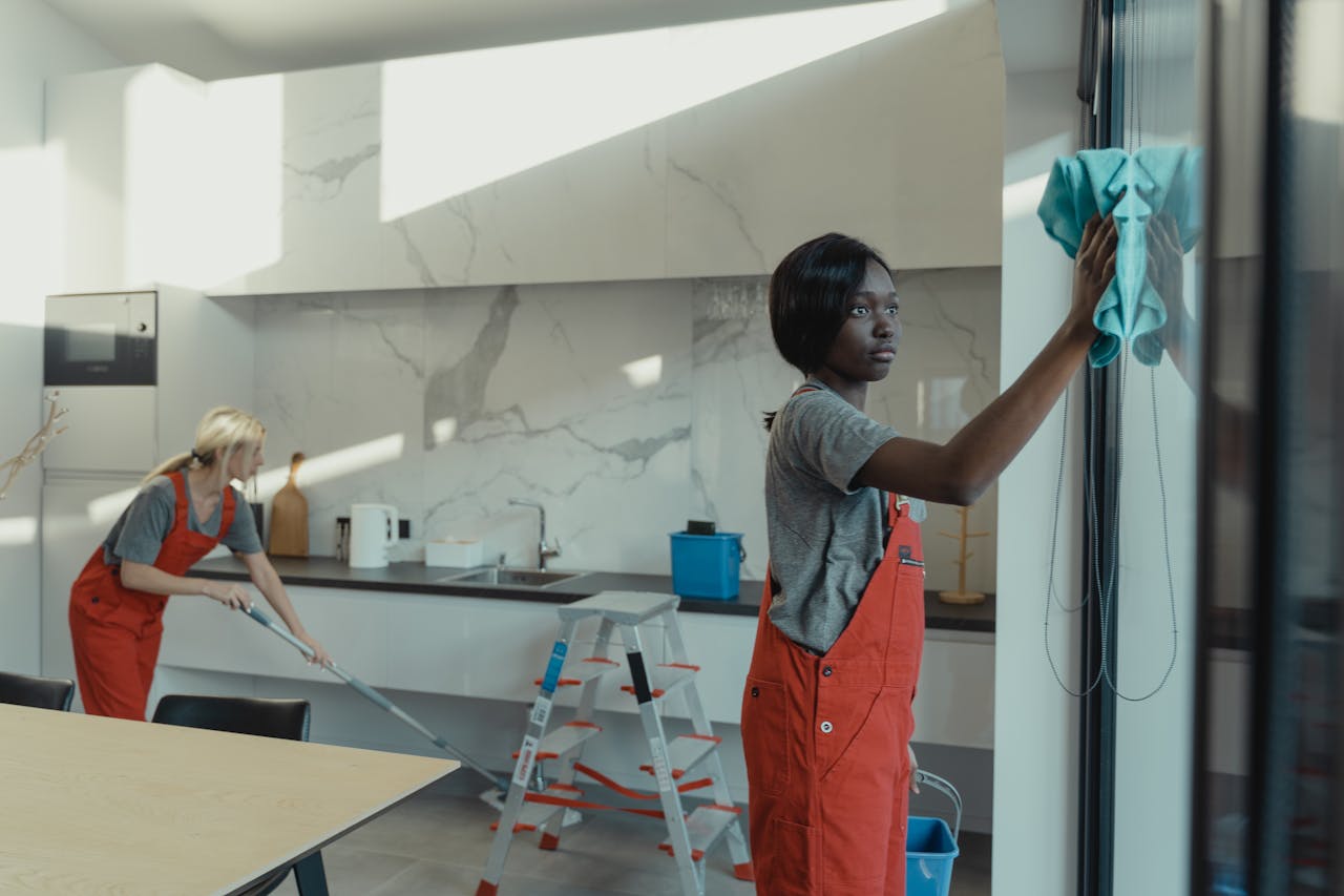 services-03 Two women cleaning a modern kitchen, wiping glass surfaces and mopping the floor.