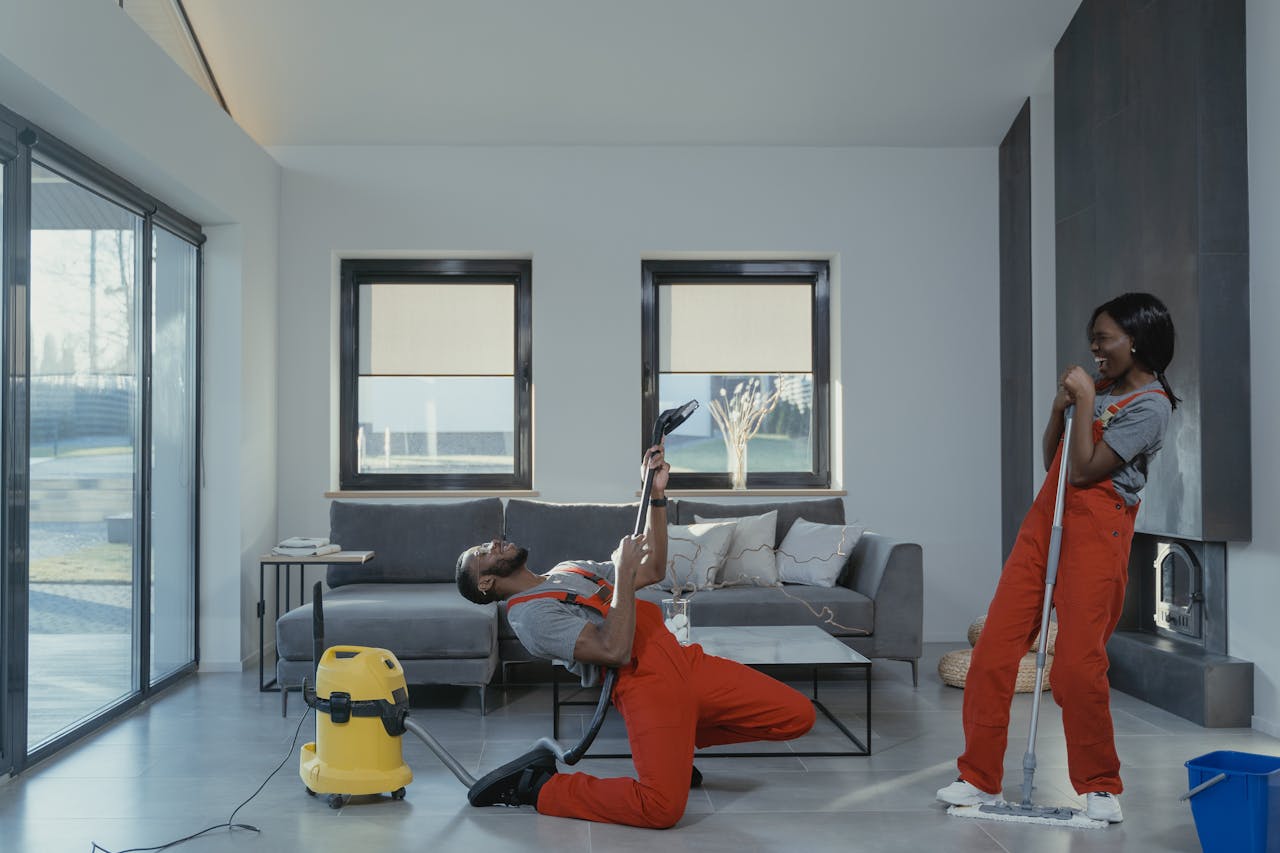 hero-img-02 Fun and engaging cleaning session with a man and woman in overalls using cleaning tools in a modern living room.